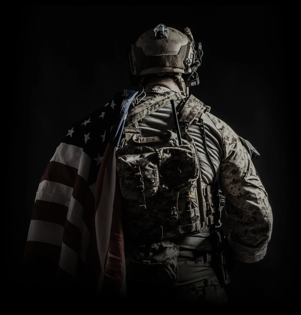 Soldier in tactical gear with an American flag draped over their shoulder, viewed from behind against a dark background.