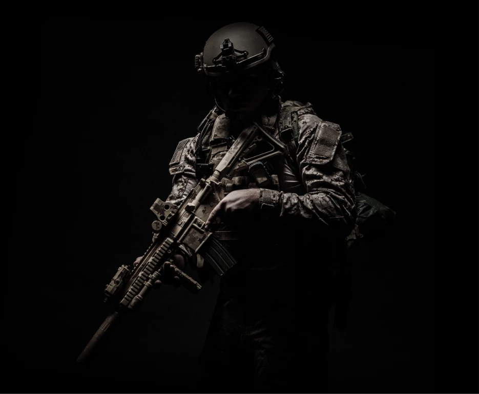 Soldier in tactical gear and helmet holding an assault rifle, partially illuminated against a dark background.