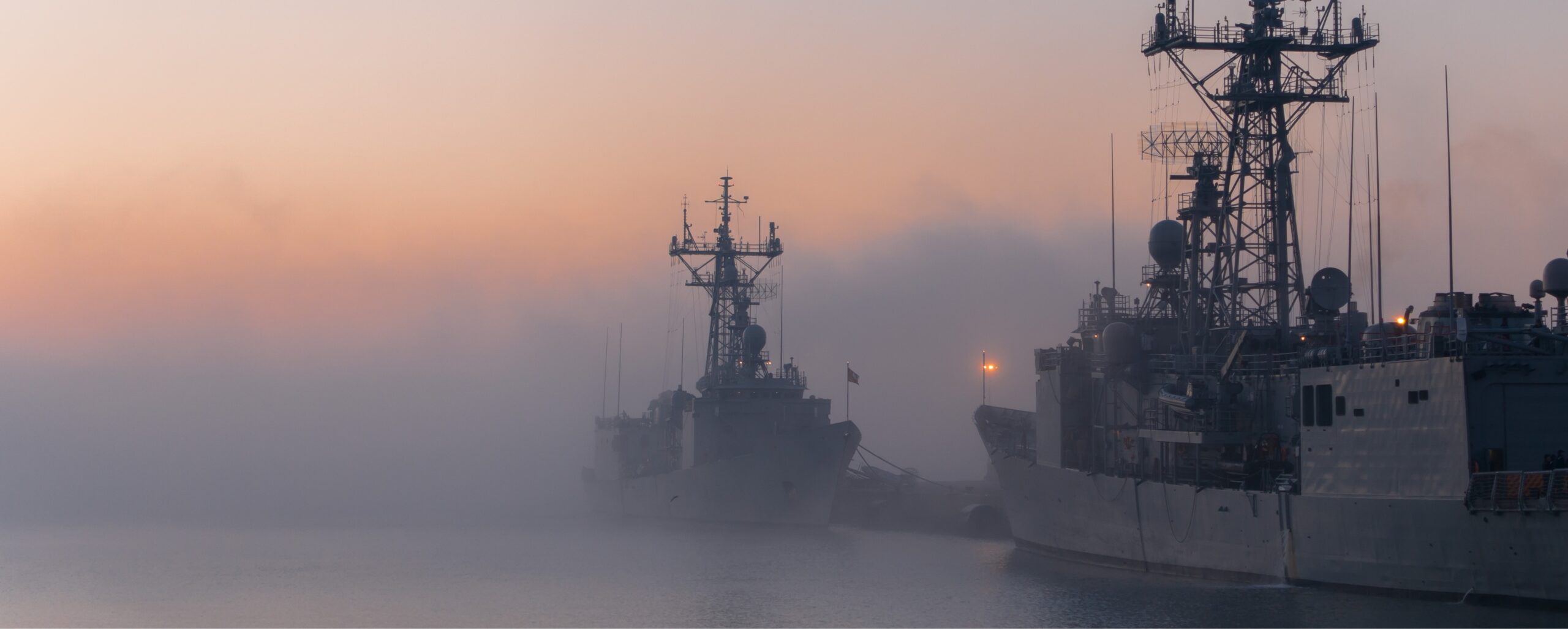 Two naval ships docked near a foggy harbor at sunset with a calm sea and a gradient sky.